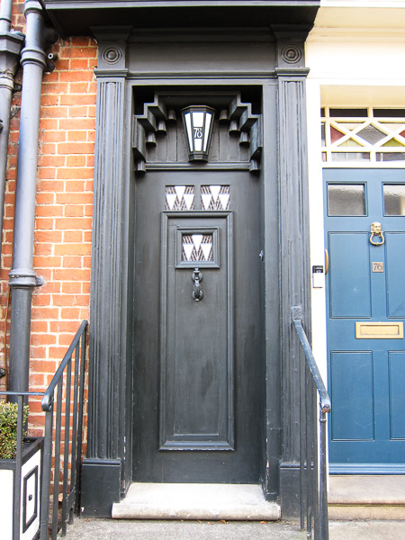a black door with black handrails in front, and one stone step.  A light in the shape of a lantern with the number 78 on it is above the door, and there are three small panels in the door with three white downwards facing triangles.