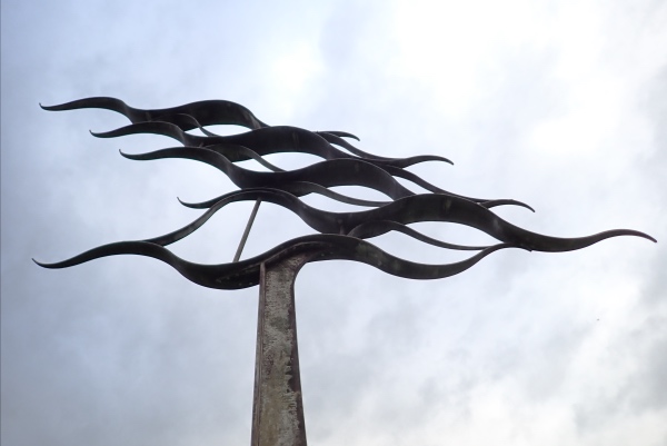 a metal sculpture against a grey sky.  There is a central pillar and various waves above it